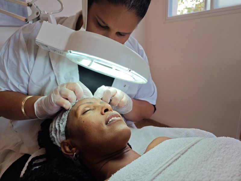 A spa professional conducts a detailed skin examination using a magnifying lamp to analyze the client's skin condition. Wearing gloves for hygiene, the specialist focuses on ensuring precision and care during this personalized skincare treatment in a bright and serene spa setting.