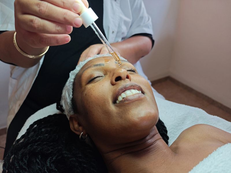 A serene moment captured during a skincare session, as a beautician carefully applies golden rejuvenating serum to the client's forehead using a dropper. The client enjoys the soothing process, enhancing skin hydration and glow.