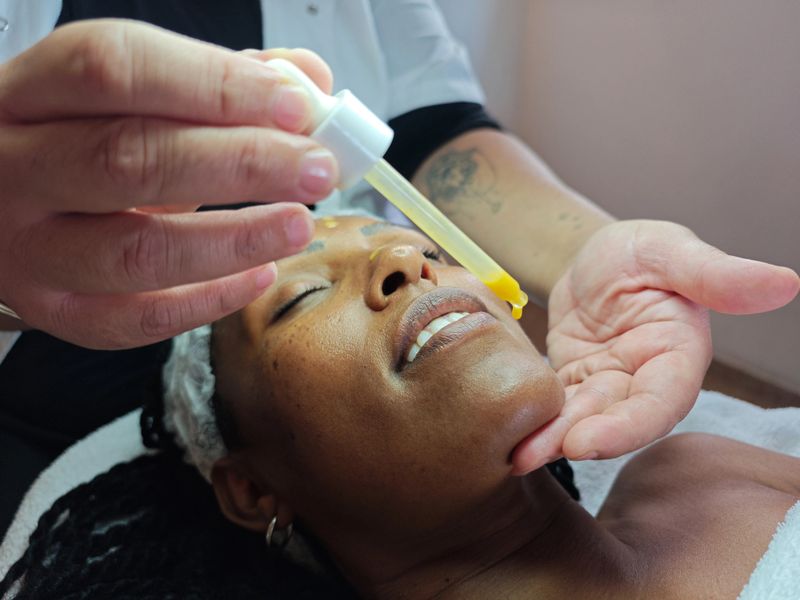A serene moment during a professional skincare session as an aesthetician applies rejuvenating facial serum to the client's forehead using a dropper. The client appears relaxed and glowing, enjoying the luxurious and soothing spa treatment in a tranquil setting.