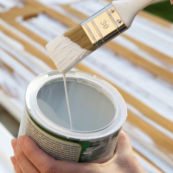 Hand dipping a paintbrush into white paint from an open can.