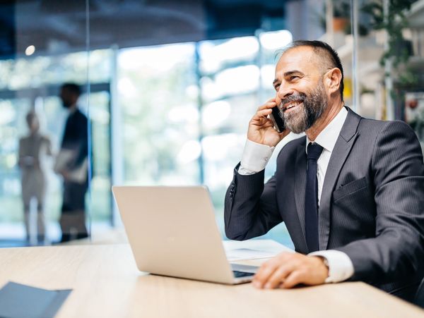 Smiling businessman talking on phone at office desk with laptop.