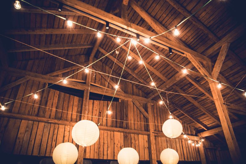 The rustic interior of a barn features wooden beams and a warm ambience created by hanging lanterns and string lights. This cozy setup is perfect for an evening gathering or celebration.
