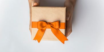 Hands holding a gift box wrapped with an orange ribbon bow.