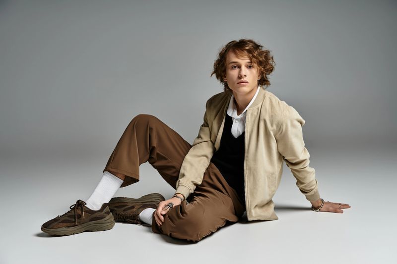A handsome young man relaxes in a chic studio, showcasing his fashionable autumn outfit with flair.