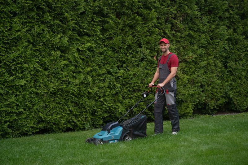 Gardener wearing a red cap and overalls mows the lawn with a modern lawnmower, working efficiently near a hedge in a well-maintained garden during a sunny day