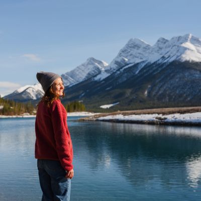 A woman smiles by a serene lake with snowy mountains in the background.