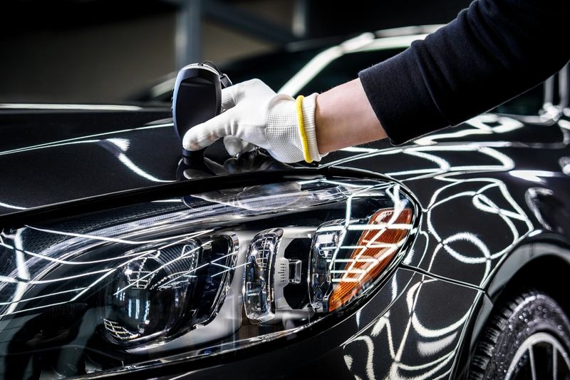 Technician measures paint thickness on car near headlight, ensuring even application and high-quality finish for automotive standards