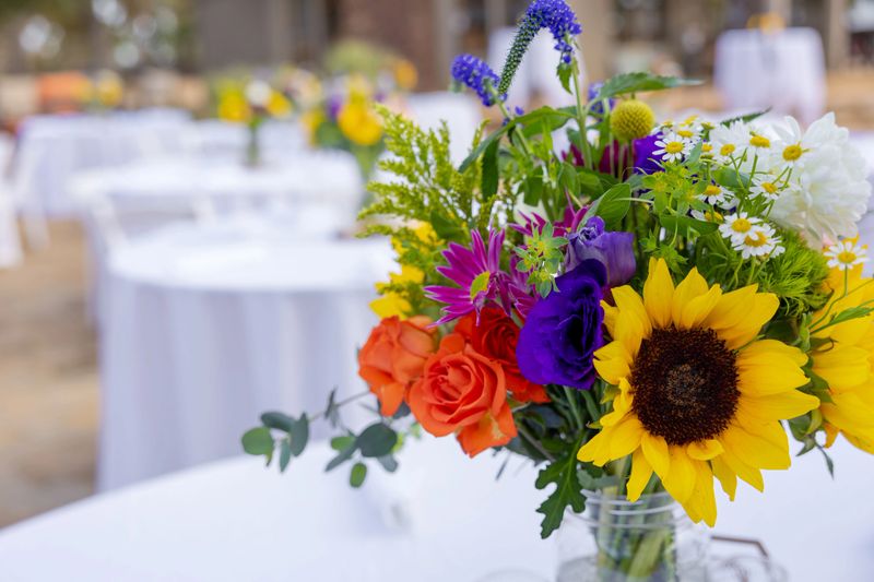 Vibrant floral centerpiece with sunflowers and roses on a white table at an outdoor event.