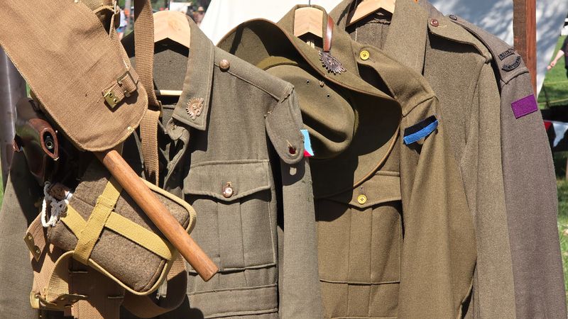A collection of vintage WWI military uniforms and equipment, including jackets with insignias, a hat, a canteen, and a shovel, all hanging on wooden hangers.