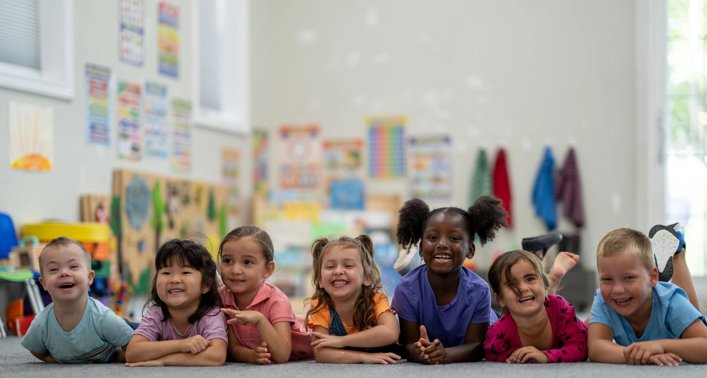 Seven diverse children lying on the floor smiling happily in a colorful classroom.