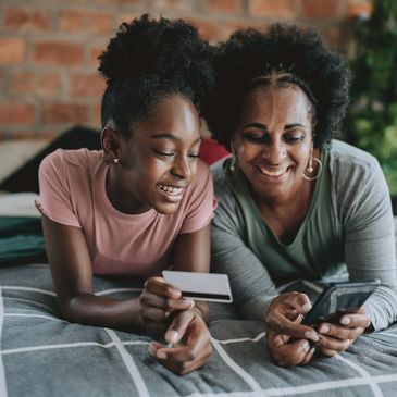 Two women shopping online together, smiling and relaxed on a bed.