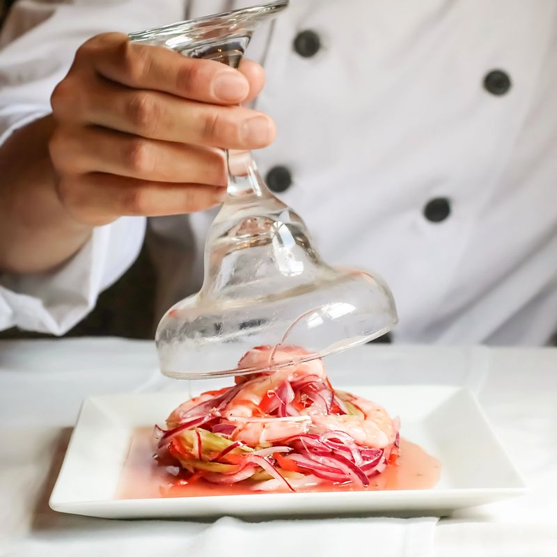 Chef is lifting a wine glass to reveal a vibrant shrimp ceviche, showcasing the dish's freshness and artful presentation