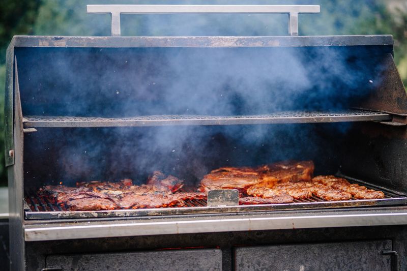 Open barbecue grill filled with a variety of meat cuts, surrounded by smoke, creating an outdoor cooking ambiance.