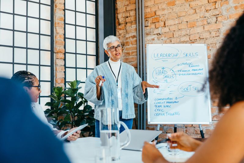 A mature businesswoman is presenting a strategy on leadership skills to engaged, diverse colleagues in a fresh and innovative office setting.