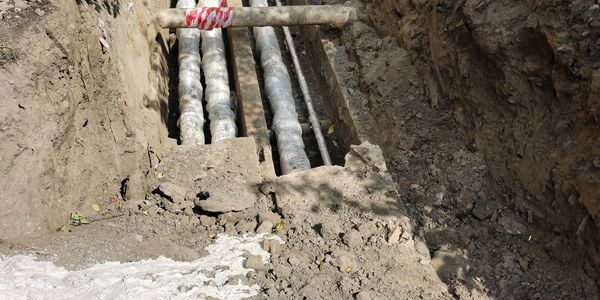 Excavated trench with insulated pipes and wooden supports near a building.
