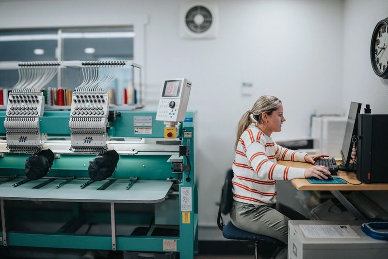 A woman who works in a manufacturing facility uses a computer to adjust settings on an industrial embroidery machine and to revise design elements being sewn onto baseball hats.