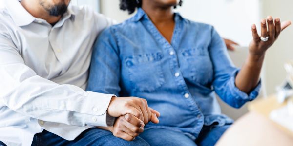 A Black couple sitting together on a couch. The man has his arm behind the woman, the woman has her 