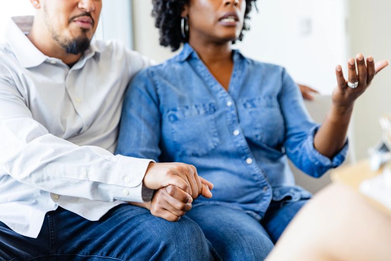 While holding her husband's hand, a worried woman talks about something with an unrecognizable counselor.