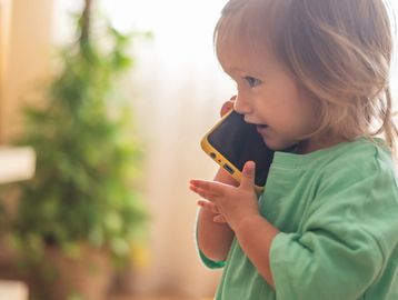 A toddler talking on a smartphone with a yellow case indoors.