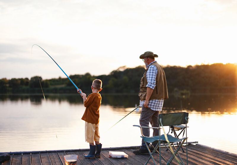 A serene scene of a grandfather and his grandson fishing together on a wooden pier as the sun rises over the tranquil lake, fostering moments of bonding and tranquility.