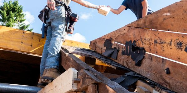 Two construction workers exchanging materials on a wooden roof structure under a clear sky.