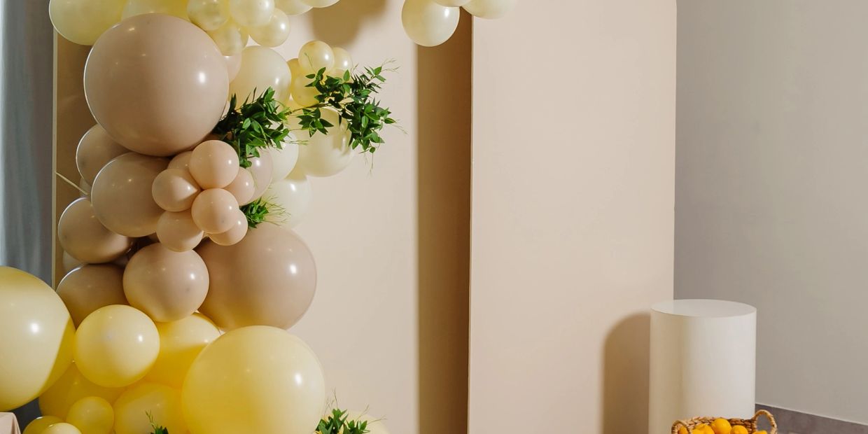 Elegant balloon arch decoration with beige and yellow balloons and green foliage accents.