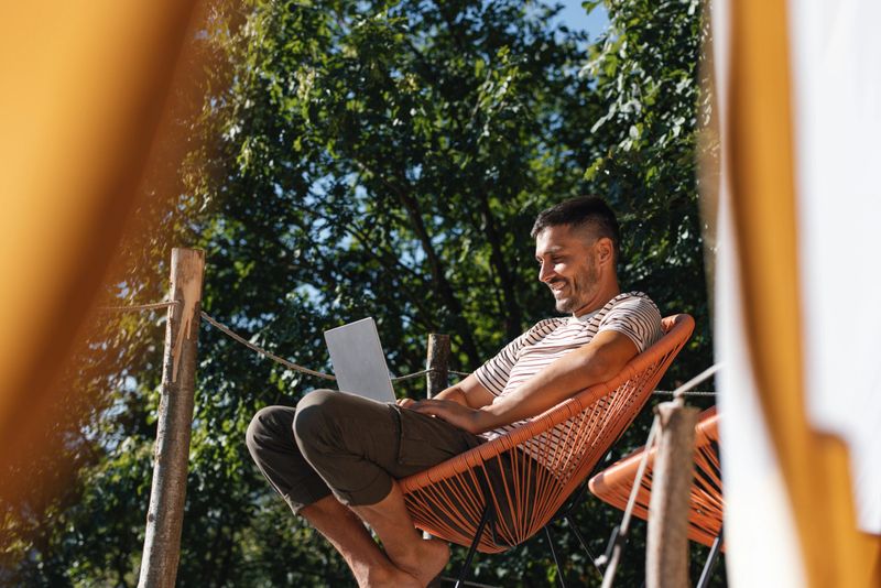 A man sits comfortably on a chair outdoors, enjoying a sunny day while working on his laptop. The peaceful scene is surrounded by lush greenery, creating a sense of relaxation and happiness.