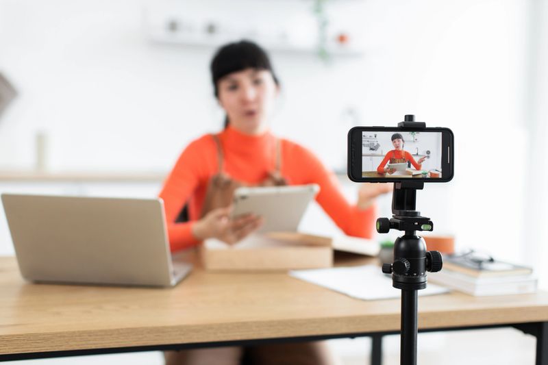 Young Caucasian female blogger unboxing new digital tablet online. Engaged influencer discussing product features while recording video content. Home workspace with modern technology setup.