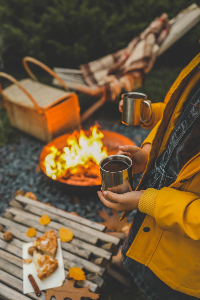 Children in warm clothing holding cups of hot drink near a bonfire in the evening. Cozy autumn outdoor scene with a fire pit, fallen leaves, and rustic snacks, capturing the essence of fall warmth.