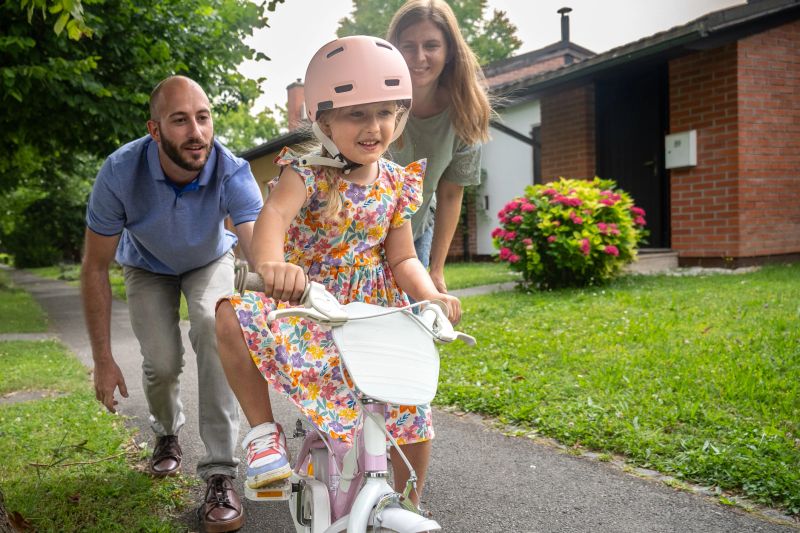 A joyful Caucasian girl learns to cycle as attentive parents support her on a sunny suburban pathway, safety helmet on and focus evident.