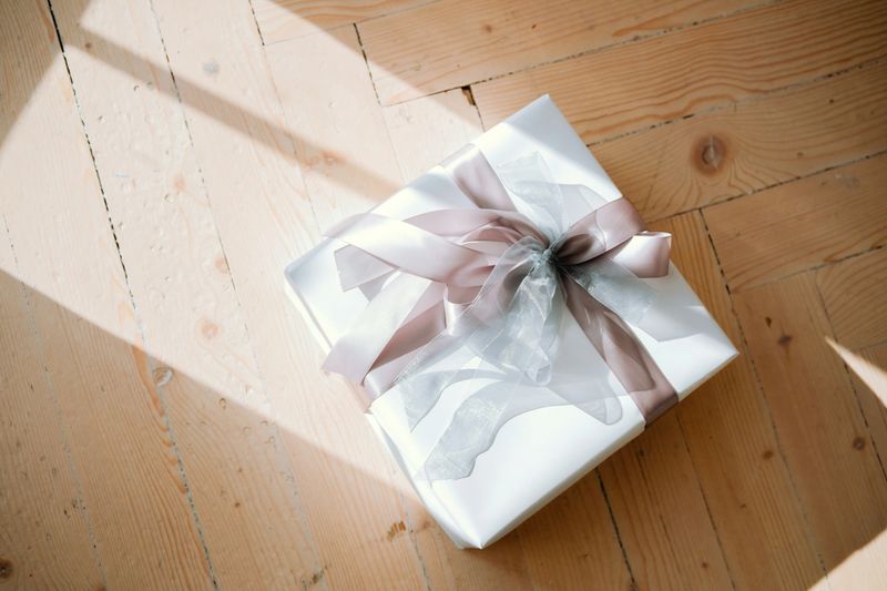 A neatly wrapped gift in soft white paper with a delicate ribbon rests on a wooden floor, illuminated by gentle sunlight streaming through a nearby window.