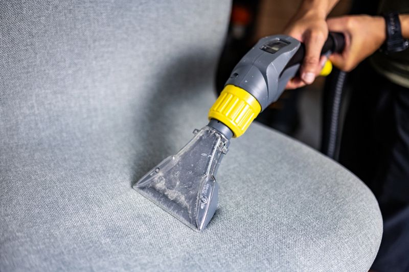 A person operates a handheld vacuum cleaner with an upholstery attachment for deep cleaning a fabric chair, focusing on hygiene and cleanliness in an indoor setting.