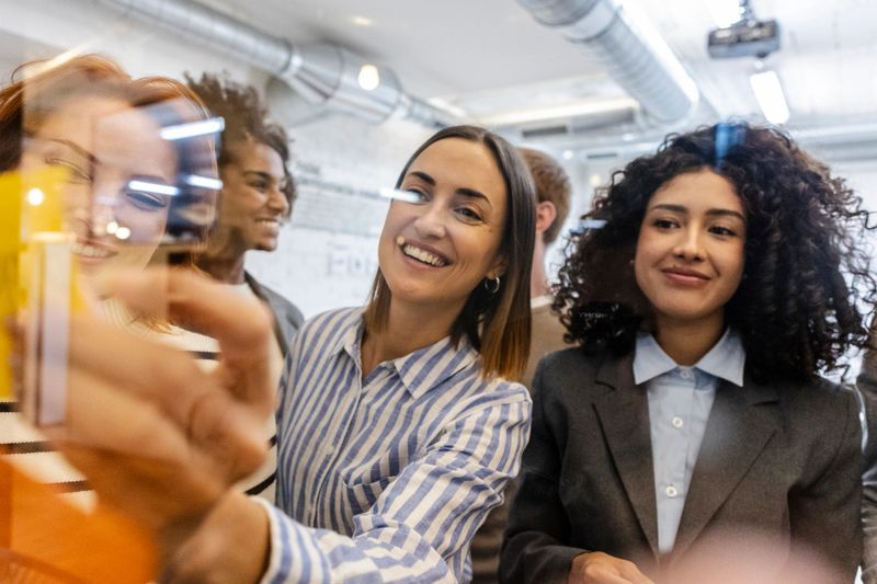 Multiracial businesswomen are smiling and collaborating on a project. Using colorful sticky notes on a glass wall in a modern coworking space. Demonstrating teamwork. Diversity. And social inclusion