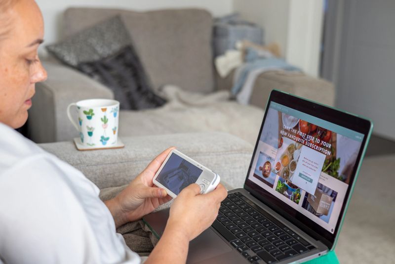 A mother sits comfortably on a sofa, enjoying a hot drink while browsing an online shopping website on her laptop. Nearby, a baby monitor screen shows her infant's room, allowing her to multitask and keep an eye on her baby while managing daily tasks.Videos are available similar to this scenario.