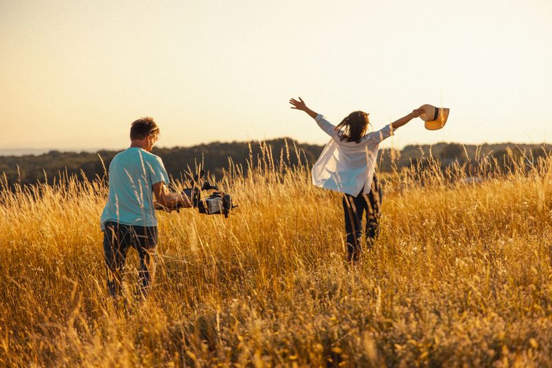A skilled cameraman records a happy Caucasian woman running through a golden field with arms raised, both clad in casual outfits, capturing a dynamic outdoor scene.