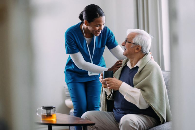 A compassionate nurse in a blue uniform helps an elderly man with a drink, creating a warm and caring atmosphere in a home setting. The scene highlights caregiving and support.