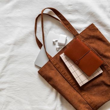 Brown tote bag with a laptop, notebooks, and wireless earbuds on a white surface.