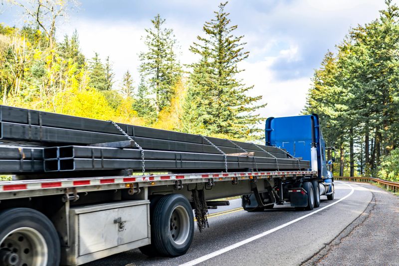 Powerful blue big rig semi truck with extended cab for truck driver rest transporting fastened by chains metal cargo on flat bed semi trailer running on the autumn road with forest in Columbia Gorge
