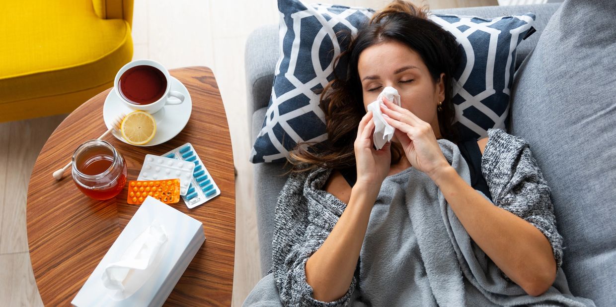 Woman resting on couch with cold medicine, tea, and tissues nearby.