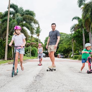 A man skateboards while three children ride scooters on a street lined with palm trees.