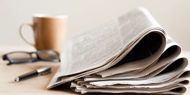 Folded newspaper on a table with a cup, glasses, and pen.