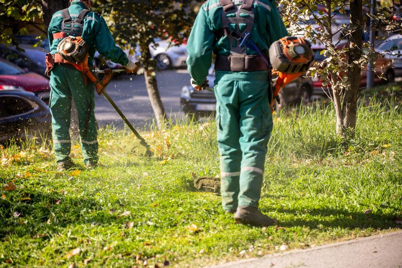 Two landscapers in green uniforms are trimming grass in a city park while enjoying a sunny day. Their focus on maintaining the landscape enhances the vibrant environment.