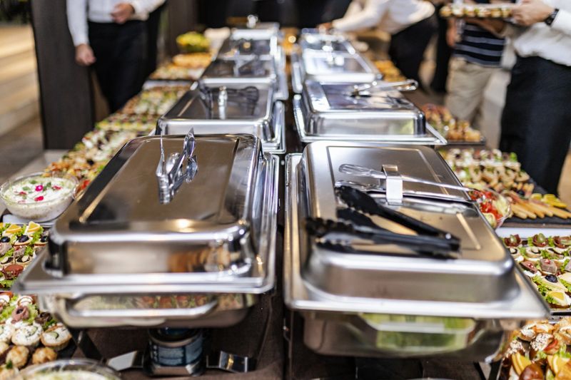 A sophisticated buffet is laid out, featuring an array of dishes including appetizers and desserts, with guests preparing to serve themselves. The atmosphere is lively and upscale.