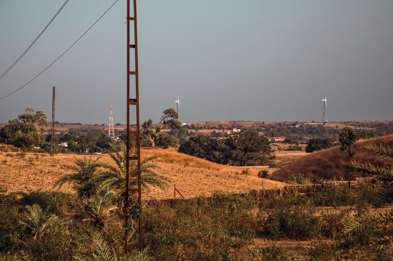 A tranquil landscape in Maharashtra features dry fields and wind turbines against a clear sky in the late afternoon. Vegetation and distant hills complete the peaceful rural setting.