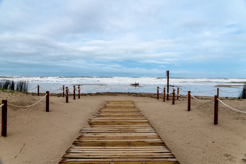 Wooden path leads from sandy beach to stormy sea, flanked by posts; driftwood and seaweed scattered on shore after storm.