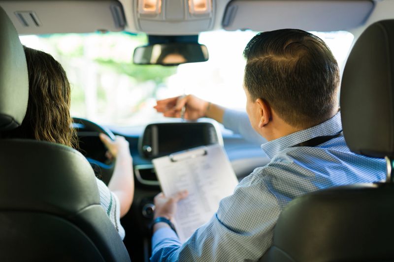 Hispanic driving instructor guiding teen student during driving test lesson, holding clipboard and giving instructions from rear view