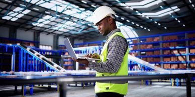 Warehouse worker in safety gear using laptop amidst storage racks.