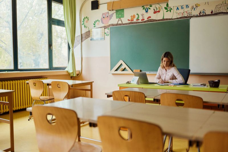 Mid adult female teacher reading and grading exam papers in the classroom. Copy space.