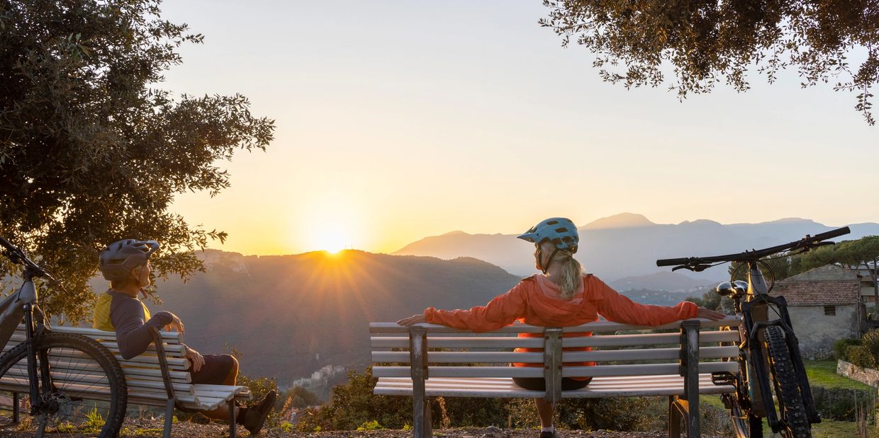 Two bicyclists sitting on benches enjoying a mountain sunset.