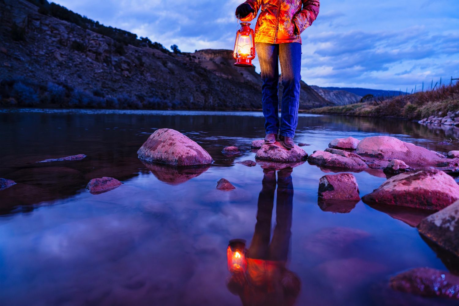Person standing on rocks by a river holding a glowing lantern at dusk.